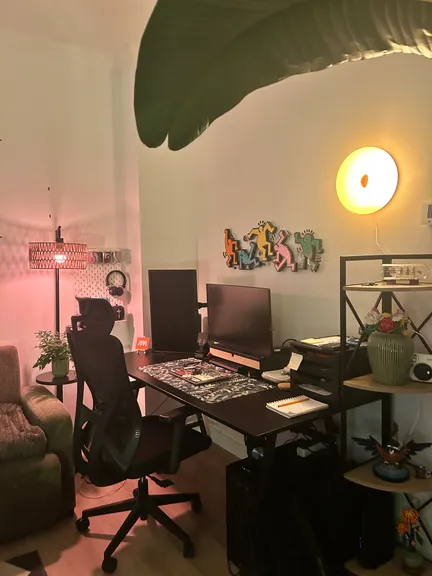 A home desk corner at night with two monitors, a mesh office chair, a paper lamp and colourful dancing figures on the wall