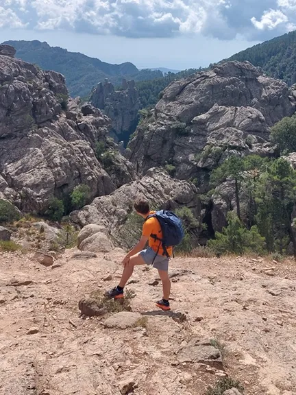 Florian in an orange shirt and a hiking backpack, standing on a rocky trail above a valley of granite spires in Corsica