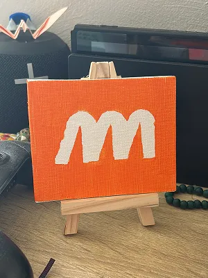 A small orange canvas on a miniature wooden easel, painted with the white M of the Modeo logo in thick brushstrokes