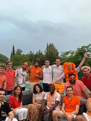 The Modeo team, about twenty people posing by a swimming pool under a grey sky, many of them dressed in orange and one in a full orange costume
