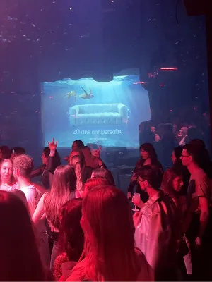 Vente-unique’s 20th anniversary party, a crowd in a dark venue lit in blue and red, facing a large screen that shows an underwater scene with a sofa and the words “20 ans anniversaire”